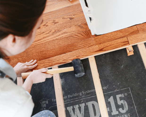 Person using a rubber mallet to install hardwood flooring over wooden floor joists and a black underlayment.