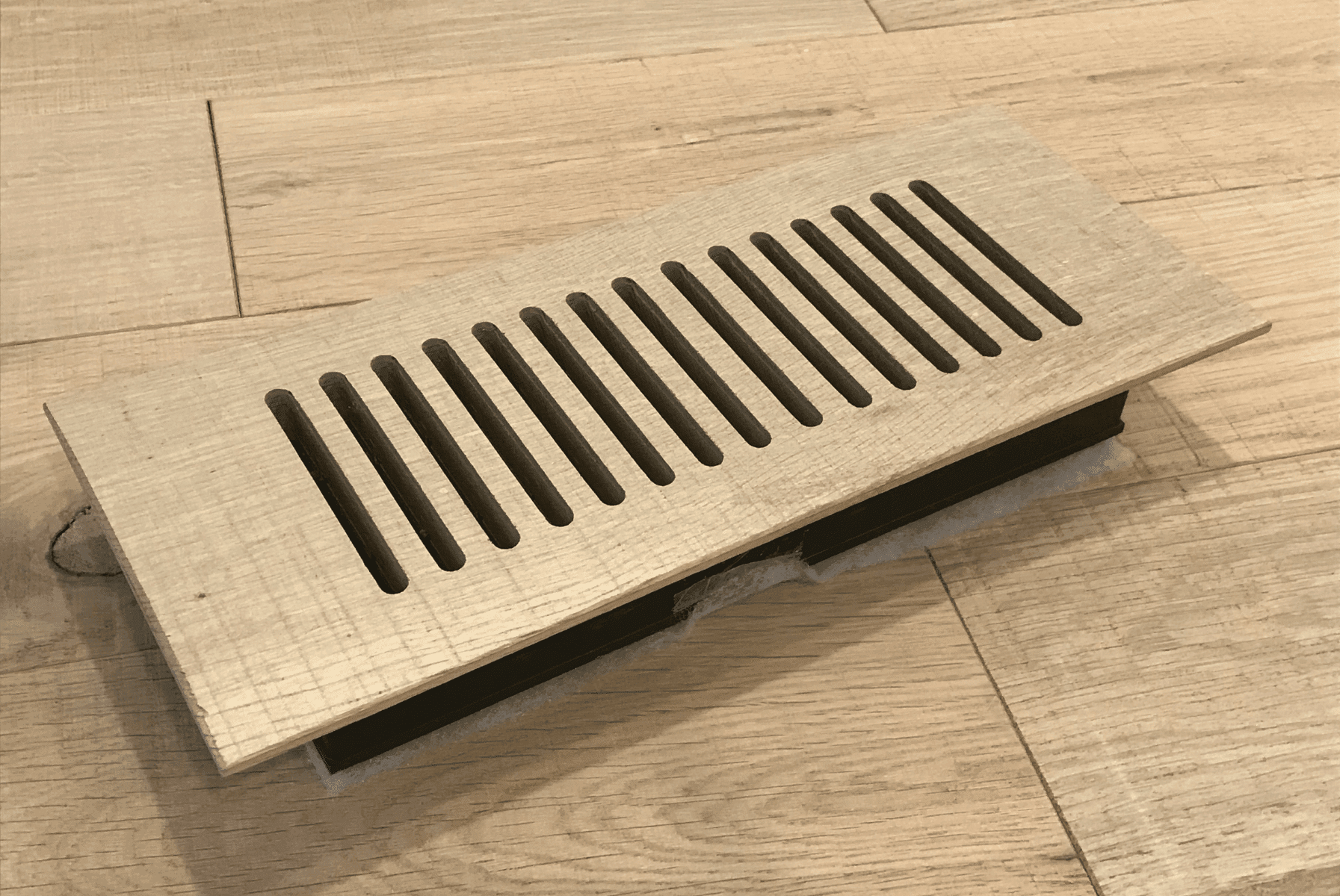 A rectangular wooden floor vent cover with vertical slats is installed in a light-colored wooden floor.