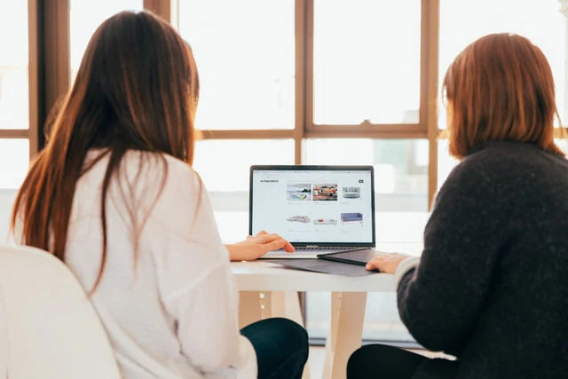 Two people sitting at a table using a laptop, looking at a screen displaying various images, in a well-lit room.